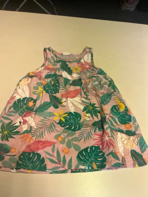 Rosa ärmlös klänning med tropiskt mönster - Söt rosa ärmlös klänning med tropiskt blommigt och bladigt mönster i grönt, gult och vitt. Lätt och luftig modell, perfekt för varma dagar. Tillverkad i mjuk bomull och har en ledig passform.