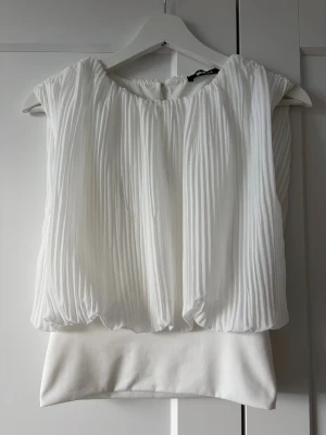 Vit plisserad blus från Gina Tricot - Snygg vit plisserad blus från Gina Tricot med rund hals och breda axlar. Blusen har ett luftigt och elegant fall med plisserat tyg upptill och slät nederdel. Perfekt för dig som vill ha en stilren och modern look.