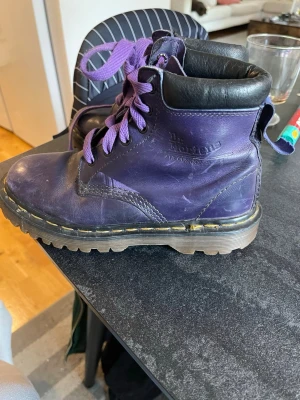 Lila Dr. Martens kängor stl 36 - Säljer ett par lila Dr. Martens kängor med svarta detaljer och lila skosnören. Klassisk grov sula med gul söm runt om och vadderad svart kant upptill. Perfekta för dig som vill sticka ut med färg på fötterna!Obs en ögla har släppt och de är smårepiga men sulorna som nya