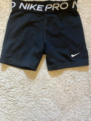 Svarta Nike Pro träningsshorts - Säljer ett par svarta Nike Pro träningsshorts med vit logga på benet och bred elastisk midja med Nike Pro-tryck. Perfekta för gym, löpning eller sport. Materialet är stretchigt och följsamt för maximal rörelsefrihet.