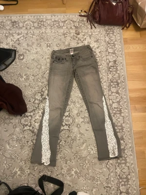Low waist Grå bootcut true religion med lace - Säljer ett par grå bootcut jeans från True Religion med vita spets längs utsidan av benen. Köpt på en second hand butik i new york. Pris kan diskuteras💕
