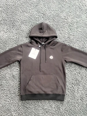 Svart hoodie från Moncler - Snygg svart hoodie från Moncler med klassisk huva och dragsnören. Har en liten Moncler-logga på bröstet och en stor magficka framtill. Tillverkad i mjukt material som känns skönt mot huden. Perfekt för dig som gillar stilrena och exklusiva plagg.