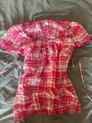Rutig rosa offshoulder blus - Söt blus i rosa och vitt rutigt mönster med offshoulder-modell och korta puffärmar. Blusen har knappar framtill, smock vid ärmslut och nederkant samt knytband i midjan för att skapa en figurnära look. Perfekt för en somrig och avslappnad look.