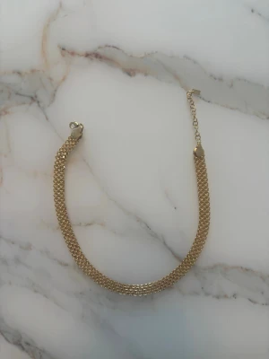 Guldigt kedjehalsband i metall - Stilrent halsband i guldtonad metall med bred, platt kedja och justerbar längd. Kedjan har ett tätt, flätat mönster som ger ett elegant och modernt intryck. Perfekt accessoar för att lyfta din outfit med en touch av guld.