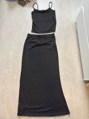 Svart glittrig croppad topp och kjol - Snygg svart croppad topp med tunna axelband och glittrigt tyg tillsammans med kjol. Perfekt för en trendig look eller till festival. Toppen och kjolens material har en lätt stretch och glittrande finish som ger extra edge. Helt oanvända. 