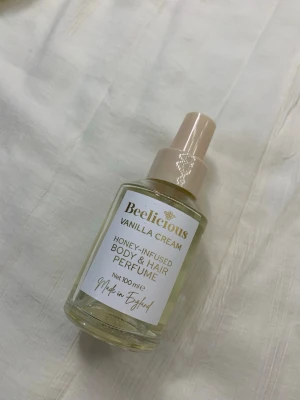 Beelicious Vanilla Cream parfym 100 ml - Fräsch parfym för kropp och hår från Beelicious i doften Vanilla Cream. Kommer i en genomskinlig glasflaska med beige spraykork och guldiga detaljer. Innehåller 100 ml och är infuserad med honung för en söt och mjuk doft.