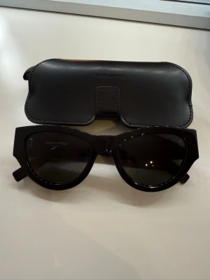 Svarta solglasögon från Saint Laurent - Snygga svarta solglasögon från Saint Laurent med bred båge och klassisk cat eye-form. Kommer med ett stilrent svart fodral med logga. Perfekta för att lyfta din look med en touch av lyx. Endast använda en gång! Inga defekter. Kvitto finns