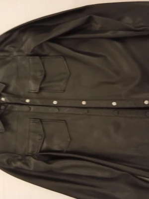 Svart skinnjacka från NAKD - Stilren svart skinnjacka från NAKD med klassisk skjortkrage och tryckknappar framtill. Två bröstfickor med lock och knappdetaljer ger en cool look. Jackan har raka ärmar och en något längre passform som passar till många olika outfits.
