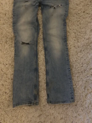 Ljusblå slitna bootcut jeans - Säljer ett par ljusblå bootcut jeans med slitningar och hål på knäna och låren. Jeansen har en tvättad look och klassisk femficksdesign. Perfekta för en avslappnad och trendig stil.