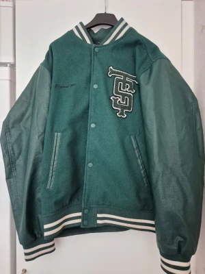 Bomberjacka - Snygg grön bomberjacka med vita detaljer och broderad logga på bröstet. Jackan har ärmar i konstläder, kropp i tyg och klassisk randig mudd vid ärmslut, krage och midja. Stängs med tryckknappar och har två fickor framtill.