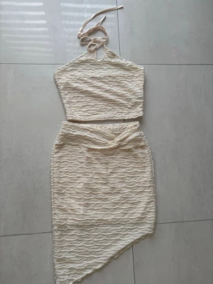 Beige spetsset med asymmetrisk kjol - Säljer ett snyggt tvådelat set i beige spets med halterneck-topp och knytning i nacken. Kjolen har en asymmetrisk skärning och knytband i midjan. Perfekt för dig som gillar trendiga och unika detaljer. Materialet är stretchigt och följsamt.