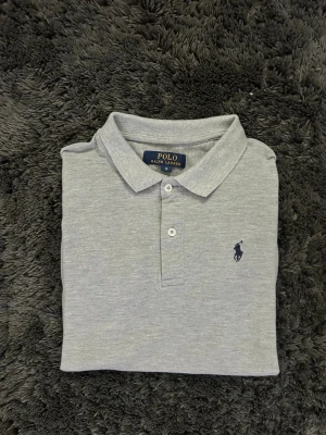 Grå Polo Ralph Lauren Piké - Klassisk grå pikétröja från Polo Ralph Lauren med korta ärmar och krage. Tröjan har två knappar framtill och den ikoniska broderade loggan på bröstet. Perfekt basic till garderoben och lätt att matcha med allt.