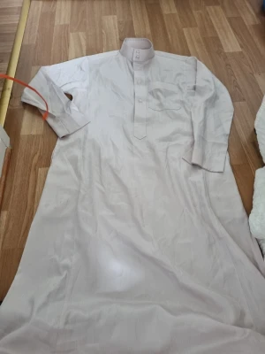 Ljusbeige lång traditionell thobe - Säljer en ljusbeige lång thobe med ståkrage och knäppning framtill. Plagget har lång ärm och en bröstficka. Perfekt för dig som gillar klassisk och stilren design. Materialet känns lätt och bekvämt.