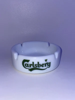Vit Carlsberg askkopp i porslin - Säljer en vit rund askkopp i porslin med Carlsberg-logga i grönt tryck på framsidan. Askkoppen har fyra urtag för cigaretter och är perfekt som samlarpryl eller dekoration.