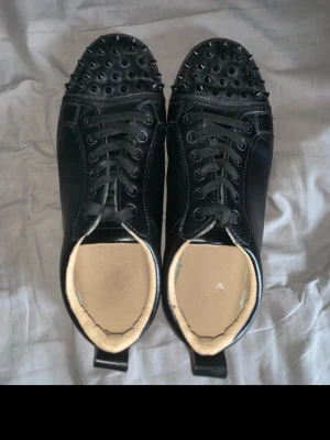 Svarta Louboutin sneakers med nitar - Svarta sneakers från Christian Louboutin med coola svarta nitar på tån och klassisk röd sula. Skorna har snörning och är gjorda i läder med en lyxig känsla. Perfekta för dig som vill sticka ut med en edgy och exklusiv stil.