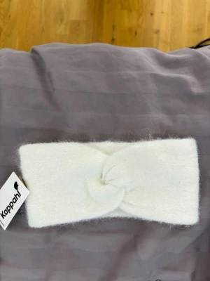 Vitt pannband från Kappahl - Mysigt vitt pannband från Kappahl i mjukt, fluffigt material. Pannbandet har en twistad knut framtill som ger en snygg detalj. Perfekt för att hålla öronen varma under kyliga dagar.