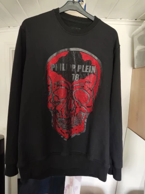 Svart Philipp Plein hoodie med dödskalle - Cool svart hoodie från Philipp Plein med stort dödskallemotiv i rött och svart framtill. Trycket är detaljerat med texten 'PHILIPP PLEIN 1978' ovanför. Tröjan har rund halsringning och är tillverkad i mjukt material, perfekt för en edgy streetstyle.