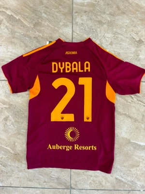 AS Roma matchtröja Dybala 21 - Snygg AS Roma matchtröja från Adidas med Dybala och nummer 21 på ryggen. Tröjan är vinröd med orange detaljer, klubbmärke på bröstet och sponsortryck. Tillverkad i lätt och ventilerande material, perfekt för fotboll eller träning.