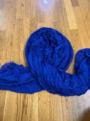 Kornblå crinklad sjal - En stor, kornblå sjal i mjukt, tunt tyg med crinklad struktur. Sjalen har fransiga kanter och är perfekt att svepa runt halsen för en cool och avslappnad look. Färgen är intensivt blå och materialet känns lätt och luftigt.