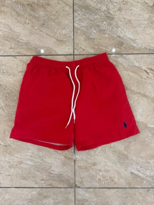 Röda badbyxor från Polo Ralph Lauren - Snygga röda badbyxor från Polo Ralph Lauren med klassisk marinblå logga broderad på benet. Modellen har elastisk midja med vit dragsko och kortare ben. Perfekta för stranden eller poolen, tillverkade i snabbtorkande material.
