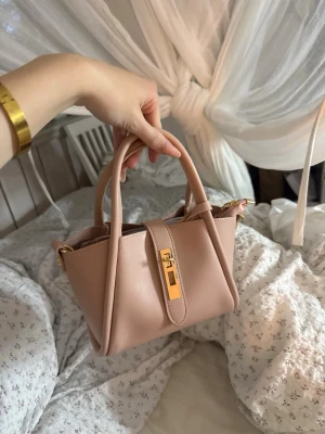 Ljusrosa handväska med gulddetaljer - Söt ljusrosa handväska i slätt konstläder med två handtag och guldfärgat spänne framtill. Väskan har en stilren och modern design med strukturerad form och detaljer i guld. Perfekt accessoar för att lyfta din outfit.