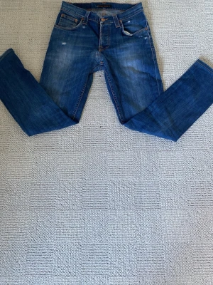 Nudie Jeans w29 - Säljer ett par klassiska blå jeans från Nudie Jeans med raka ben och femficksmodell. Jeansen har snygga slitningar framtill och detaljerade bakfickor med Nudie-loggan. Tillverkade i denim av bomull och har orangea sömmar. De har blivit lagade av Nudie själva innan därför säljs billigt.
