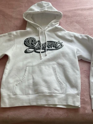 Vit hoodie från Element med tryck - Snygg vit hoodie från Element med stort logotryck på bröstet. Klassisk känguruficka framtill och dragsko i huvan. Perfekt för en avslappnad look! Storlek L på lappen men passar mer som en S enligt mig. 