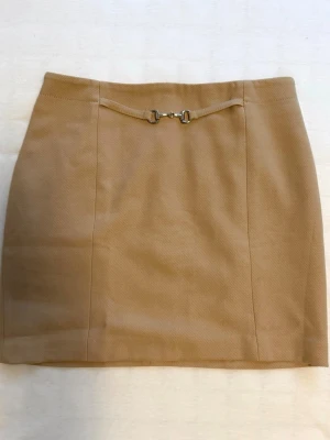 Beige kortkjol med gulddetalj - Stilren beige kortkjol med snygg guldfärgad detalj framtill. Kjolen har en rak passform och är perfekt för dig som gillar en klassisk och trendig look. Materialet känns mjukt och har en lätt struktur 🤍
