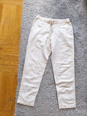 Beiga Uniqlo linnebyxor med dragsko - Snygga beiga Uniqlo linnebyxor med avslappnad passform och dragsko i midjan. Byxorna har raka ben och sidofickor, perfekta för en chill och stilren look. Tillverkade i ett lätt och luftigt linnematerial som passar bra till varmare dagar.