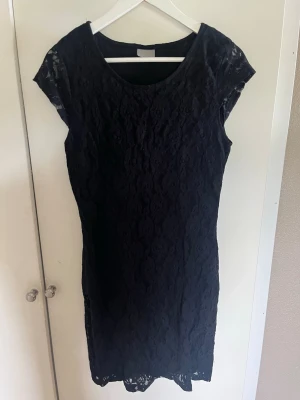 Svart spetsklänning med kort ärm - En svart, figurnära spetsklänning med korta ärmar och rund halsringning. Klänningen har ett elegant spetsmönster över hela plagget och är i kort modell. Perfekt för dig som gillar klassisk och stilren design.