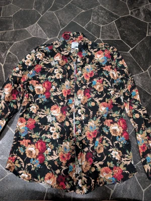 Blommig skjorta från Clubs and Spades stl L - Snygg långärmad skjorta från Clubs and Spades med färgstarkt blommigt mönster i rött, beige, blått och grönt på svart botten. Skjortan har klassisk krage och knappar framtill. Perfekt för dig som vill sticka ut med en unik stil.