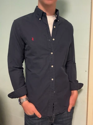Ralph Lauren marinblå skjorta – M slim fit - Säljer en klassisk och stilren marinblå skjorta från Ralph Lauren i slim fit, storlek M slimfit. Perfekt för både vardag och finare tillfällen. I mycket gott skick. Har du frågor? Tveka inte att höra av dig. Allt gott från Deluxe garderoben ⭐️  