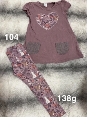 Lila set med hjärta - Sött tvådelat set med lila tunika och matchande leggings. Tunikan har ett hjärta med blommigt mönster på bröstet och två fickor framtill. Leggingsen har ett all-over tryck med blommor i lila, rosa och bruna toner. Perfekt för dig som gillar gulliga detaljer!🌸 Fint använt skick. Storlek 104