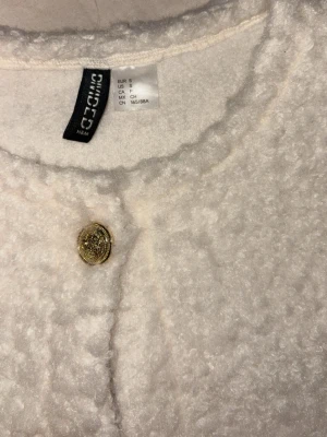 Vit bouclékofta från H&M med guldknappar - Säljer en vit bouclékofta från H&M med rund halsringning och fyra dekorativa guldknappar framtill. Koftan har lång ärm och är gjord i ett fluffigt, mjukt material som ger en lyxig känsla. Perfekt för dig som vill ha en trendig och elegant look. Köpes via Swish eller i appen men då måste ni skriva innan ☺️