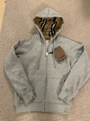 Grå zip-hoodie från Burberry - Snygg grå zip-hoodie från Burberry med klassiskt rutigt foder i huvan och dragsko. Hoodien har ribbade muddar, fickor framtill och dragkedja. Perfekt för dig som gillar stilrena plagg med exklusiva detaljer.