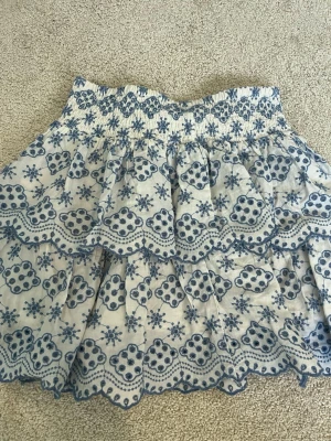 Vit och blå broderad vorlang kjol från Lindex - Supersöt kort kjol från Lindex i vitt med blått broderat mönster och volangkant. Kjolen har smock i midjan och är gjord i bomull, perfekt för en fräsch och somrig look. 