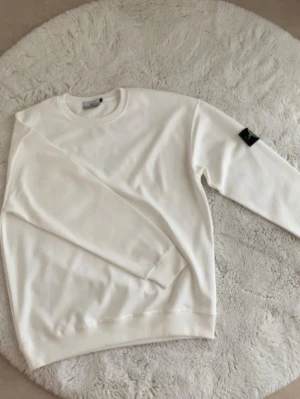 Vit sweatshirt från Stone Island - Vit sweatshirt från Stone Island med klassisk rund hals och ribbade muddar. Tröjan har en svart logotyp-patch på ena ärmen som ger en snygg streetwear-vibe. Perfekt för dig som gillar stilrena och trendiga plagg.