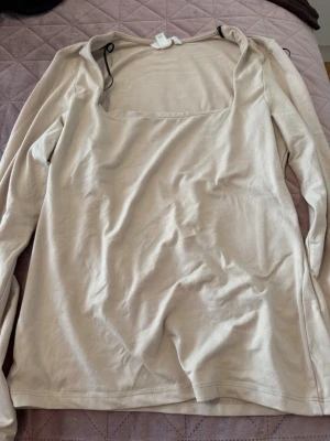 Beige långärmad topp med fyrkantig ringning - Snygg beige långärmad topp med fyrkantig ringning och enkel design. Tillverkad i mjukt bomullsmaterial som känns skönt mot huden. Perfekt att styla med jeans eller kjol för en clean look.