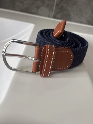 Mörkblått flätat bälte med skinn - Snyggt mörkblått flätat bälte med bruna skinn-detaljer och silverfärgat spänne. Perfekt accessoar för att lyfta din outfit med en klassisk touch. Passar till både jeans och chinos.