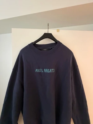 Mörkblå sweatshirt Axel Arigato - Mörkblå sweatshirt från Axel Arigato med tryckt logga i ljusblått på bröstet. Oversized passform med rund halsringning och långa ärmar. Perfekt för en chill och stilren look.