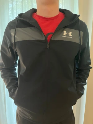 Under armour jacka svart - Svart vindjacka från Under Armour med grått parti över bröstet och vit logga. Jackan har dragkedja framtill, huva med dragsko och är tillverkad i ett lätt och smidigt material. Perfekt för en sportig och avslappnad stil. Den har dock ett hål i högra fickan. Men det är bara första lagret av tyg så fickan funkar fortfarande. Storlek:s pris kan diskuteras 