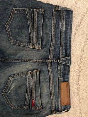 Blå ONLY bootcut jeans  - Säljer ett par blå bootcut jeans från ONLY med snygga slitningar och kontrastsömmar. Jag säljer för att de tyvärr är för stora för mig. Köpte ifrån plick innan men har aldrig använt dem. Innebenslängden är 71 cm och midjemåttet tvärs över är 41 cm.