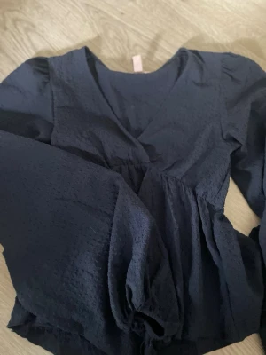 Mörkblå blus med struktur från Nelly - Mörkblå blus från Nelly med v-ringning och långärmad design. Blusen har en lätt strukturerad yta och voluminösa ärmar med resår vid ärmslut. Perfekt för dig som gillar en stilren men ändå trendig look. Helt ny använd 1 gång 