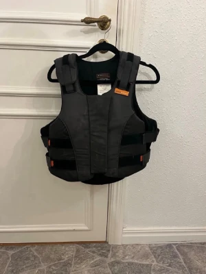 Svart säkerhetsväst från Airowear - Svart säkerhetsväst från Airowear med justerbara kardborreband i sidor och axlar. Västen har orange detaljer och är tillverkad i slitstarkt material, perfekt för dig som vill ha extra skydd vid ridning eller sport. Märkt med BETA 2009 Level 3 för hög säkerhetsstandard.