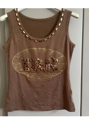 Brun linnetopp med pärldetaljer - Snygg brun linnetopp med pärldetaljer runt halsringningen och ett dekorativt mönster framtill i guld och pärlor. Toppen har ärmlös design och rund hals. Perfekt för dig som gillar unika detaljer och en avslappnad men cool stil.