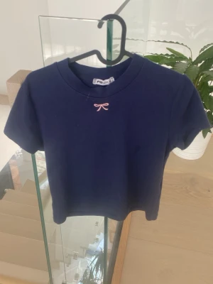 Mörkblå t-shirt med rosett - Mörkblå t-shirt med korta ärmar och en liten rosa rosett broderad vid halsringningen. Modellen är croppad och har en klassisk rund hals. Perfekt för en trendig och enkel look.