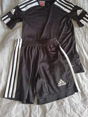 Svart Adidas träningsset med ränder - Säljer ett svart träningsset från Adidas med klassiska vita ränder på både t-shirt och shorts. Setet har tryckt Adidas-logga på ärm och ben, samt elastisk midja på shortsen. Materialet är lätt och andas, perfekt för träning eller sport. Tröjan är mer ny än shortsen. Men fungerar fint. Tvättat med flytande tvättmedel mild doft. Inga djur i hemmet. 