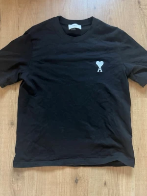 Svart t-shirt från Ami Paris - Svart t-shirt från Ami Paris med klassisk passform och korta ärmar. På bröstet finns ett vitt broderat hjärta med bokstaven A. Tillverkad i mjuk bomull och perfekt för en stilren look. Pris kan diskuteras vid snabb affär.