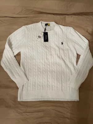 Vit kabelstickad tröja Polo Ralph Lauren - Vit kabelstickad tröja från Polo Ralph Lauren med rund halsringning och klassisk broderad logga på bröstet. Tröjan har långa ärmar och ribbade muddar vid ärmslut och nederkant. Perfekt för en stilren look.