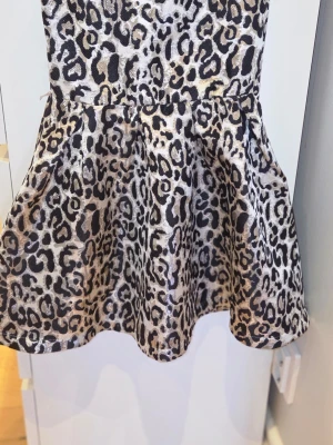 Leopardmönstrad kort klänning - Cool kort klänning med leopardmönster i beige, svart och grå toner. Klänningen har en utsvängd kjol och är ärmlös, perfekt för dig som vill sticka ut. Materialet känns som polyester och har en lätt glans.